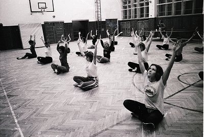 Group fitness class in a gymnasium, likely 1970s–1980s. Participants in coordinated athletic wear perform arm-raising exercis...