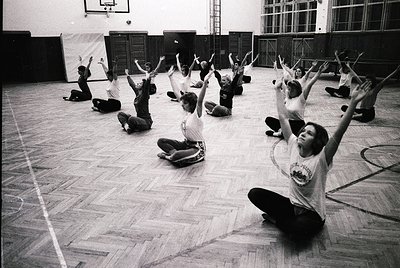 Group exercise class in a gymnasium, mid-20th century. Participants in coordinated athletic wear perform synchronized arm rai...