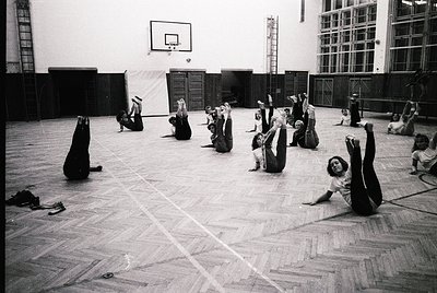 Indoor gymnasium scene with 15+ individuals in dynamic stretching poses, likely a fitness or yoga class. Wooden flooring, bas...
