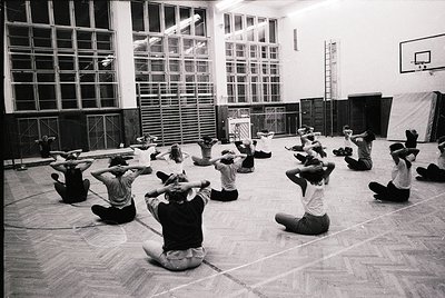 Group fitness session in a large, institutional gymnasium, likely from the 1970s. Participants perform synchronized arm stret...