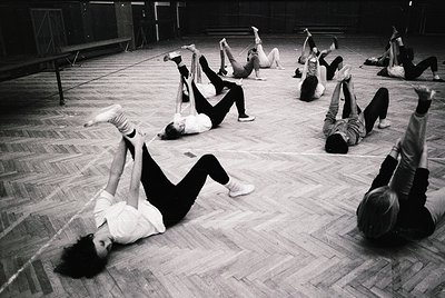 Group performing floor exercises in a spacious, wood-paneled studio—likely a 1960s–1970s dance or gymnastics class. Dynamic p...