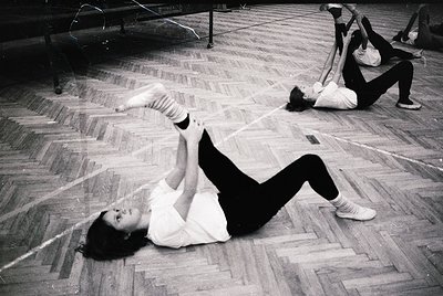 Black-and-white gym scene featuring two individuals performing floor exercises in a wooden-floored hall, likely from the 1960...