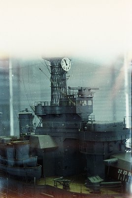 Mid-20th century naval vessel with prominent clock tower and anti-aircraft mounts, likely a WWII-era battleship. Rust and wea...