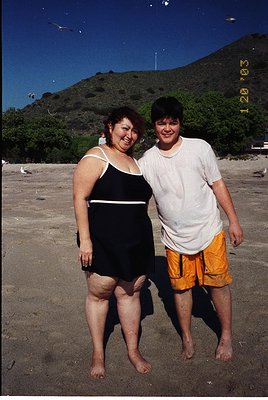 Vintage beach portrait of two individuals on a rocky shore, framed by lush green hills under clear skies. Woman in black swim...