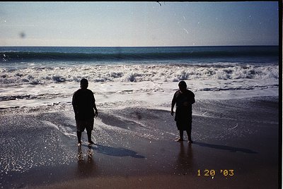 Two figures stand on a wet beach shore, facing crashing waves under a clear sky. Mid-2000s coastal scene with vintage film gr...
