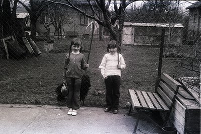 Two children pose outdoors in a residential backyard, likely mid-20th century. The girl on the left holds a broom and a bag, ...