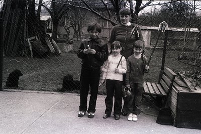 Family portrait in a residential courtyard, likely 1970s–1980s. Four individuals pose outdoors near a wooden bench and rustic...