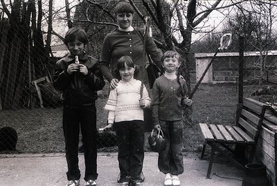 Family portrait in a park setting, likely 1970s–1980s. Three children and an adult woman pose outdoors near a bench and chain...
