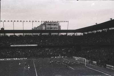 Vintage black-and-white stadium shot showing a packed arena with tiered seating and a sparse field. Visible signage includes ...