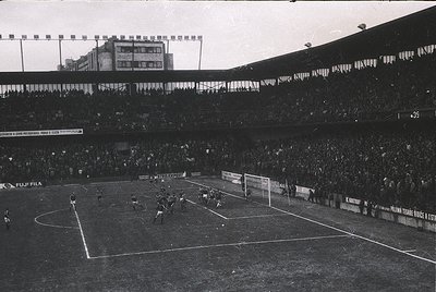 Black-and-white stadium shot capturing a 1970s European football match. Crowded stands with dense seating, visible advertisem...
