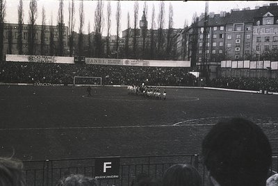Black-and-white photo of a packed stadium during a 1960s football match. Crowds line the perimeter, with visible banners adve...