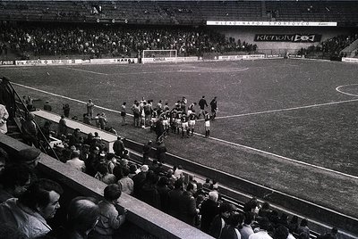 Black-and-white shot of a mid-20th century football match at a packed stadium, likely Eastern Bloc era. Players in white kits...