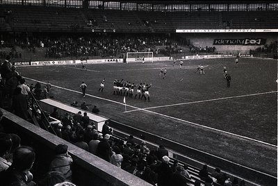 Black-and-white shot of a packed stadium during a mid-20th-century football match, likely Eastern Bloc era. Players in white ...