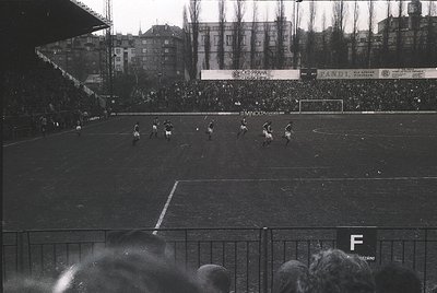 Vintage black-and-white soccer match in a packed stadium, featuring mid-action players on a grass field. Advertising banners ...