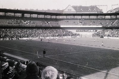 Vintage black-and-white shot of a packed stadium during a football match, likely 1960s–1970s. Multi-tiered stands filled with...