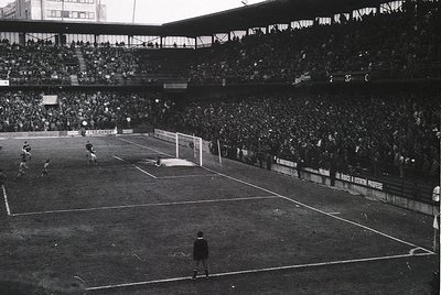 Black-and-white shot of a packed stadium during a mid-20th-century football match, likely 1950s–1960s. Crowds fill tiered sta...