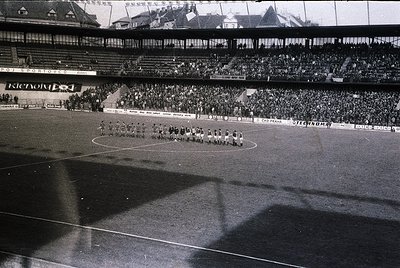 Black-and-white stadium shot showing a pre-match ceremony with teams lined up on a wet pitch. Multi-tiered seating filled wit...