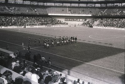 Black-and-white stadium shot showing pre-match team lineups on a grass pitch, flanked by tiered seating filled with spectator...