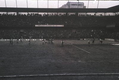 Black-and-white soccer match in a packed stadium, likely 1960s–1970s. Players in white and dark jerseys compete on a muddy fi...