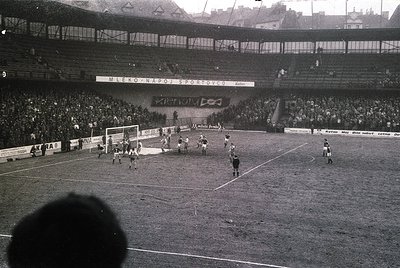 Black-and-white soccer match at a packed stadium, featuring a mid-action play near the goal. Crowds fill tiered stands with b...