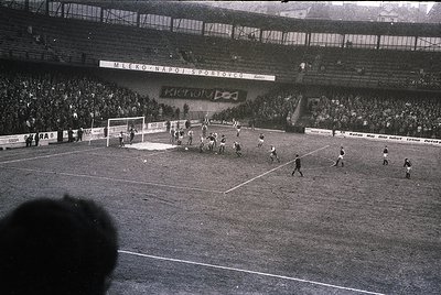 Black-and-white soccer match in a packed stadium, featuring a crowded stands with spectators and banners reading "Mesto Naro ...