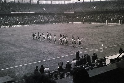 Black-and-white shot of a mid-20th-century football match, likely 1950s–1960s. Players in dark uniforms line up for a corner ...