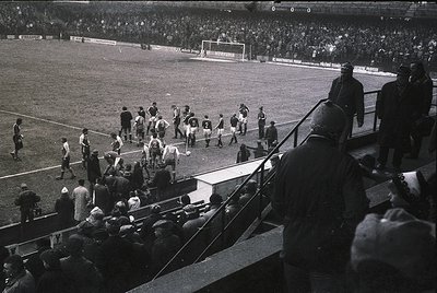 Black-and-white stadium shot showing pre-match team huddles on grass, likely 1950s–1960s. Players in white and dark striped j...