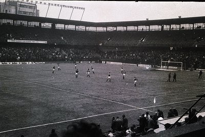 Black-and-white stadium shot featuring a mid-20th century football match. Players in dark uniforms chase the ball on a grass ...