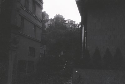 Black-and-white alleyway framed by tall, weathered buildings with exposed brick and concrete. Narrow pathway leads uphill to ...