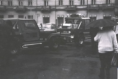 Vintage black-and-white street scene featuring a 1960s-era Jeep CJ-5 and other classic vehicles in a wet urban plaza. Multi-s...