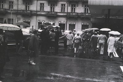 Crowded urban street scene featuring horse-drawn carriages and pedestrians under umbrellas, likely mid-20th century. Multi-st...