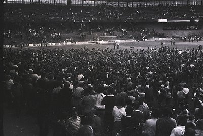 Black-and-white shot of a packed stadium during a football match, likely mid-20th century. Crowd fills stands, with visible s...
