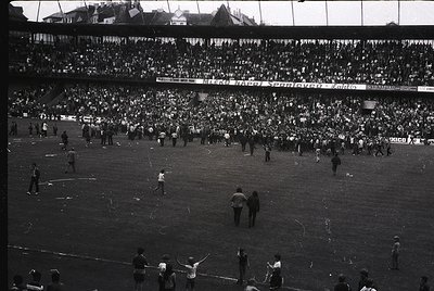 Black-and-white stadium shot showing chaotic post-match scene with players and officials on field amid dense crowd. Advertisi...