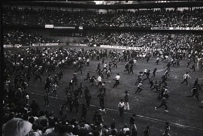 Chaotic stadium scene with players and fans flooding the field during a match. Mid-20th century stadium architecture with tie...