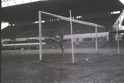 Empty mid-20th century stadium goalpost with netting damage, likely post-match. Concrete stands and sparse spectators in back...
