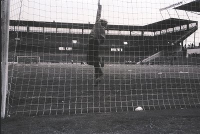 Black-and-white shot of a goalkeeper diving to block a soccer ball, mid-action. Empty stadium seats in background suggest a l...
