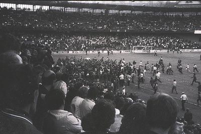 Vintage black-and-white stadium shot capturing a chaotic post-match scene, likely from the 1960s–70s. Crowd surges onto the f...