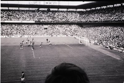 Black-and-white stadium shot captures a packed 1960s-era football match. Players in dark uniforms clash near the goal, while ...