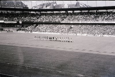 Black-and-white stadium photo showing a pre-game formation of a soccer team on a wet pitch, with a packed multi-tiered stand....