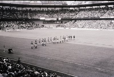 Black-and-white stadium photo showing pre-match team walkout on a grass pitch. Players in striped jerseys and shorts line up ...