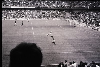Black-and-white shot of a packed stadium during a mid-20th-century football match. A player in mid-air executing a header nea...