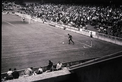Black-and-white shot of a packed stadium during a soccer match, likely 1960s–1970s. Player in motion near the center circle, ...