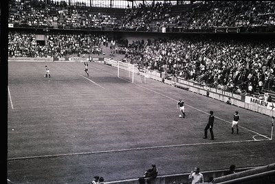 Black-and-white stadium shot capturing a 1960s–1970s football match. Crowded stands filled with spectators, visible branding ...
