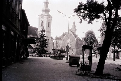 Black-and-white street scene featuring **1960s-70s European urban architecture**. Prominent **baroque-style church tower** wi...