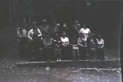 Black-and-white group portrait of 12 individuals seated on a low stone wall, likely mid-20th century. Formal attire includes ...