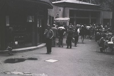 Crowded urban street scene featuring a mix of pedestrians, including a man in a suit and hat carrying a bag, near a small caf...