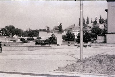 Monumental statue atop pedestal in urban plaza, flanked by low-lying Soviet-style architecture. Prominent tree-lined street w...