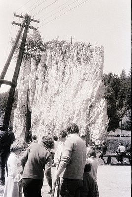 Mid-20th century group of people observing a large, weathered rock formation with a cross carved at its summit. Clothing sugg...