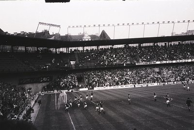 Black-and-white shot of a packed stadium during a mid-20th-century football match, likely 1950s–1960s. Crowds fill tiered sta...