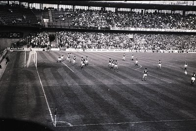 Vintage black-and-white soccer match in a packed stadium, featuring two teams in white and dark uniforms. Mid-action play nea...
